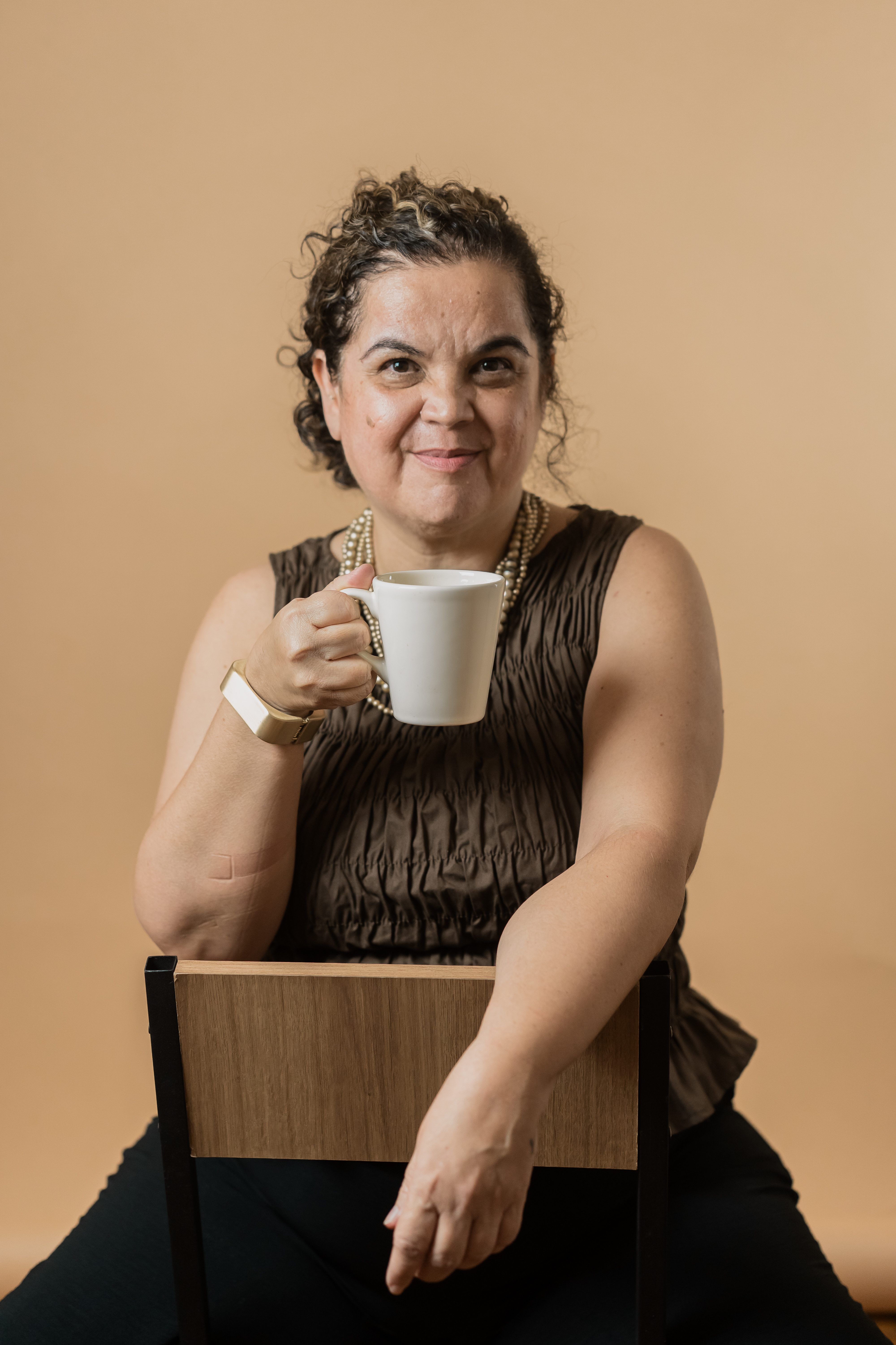 Mulher segurando uma caneca branca, sorrindo, vestindo uma blusa marrom e colar de pérolas, sentada em uma cadeira com fundo laranja.